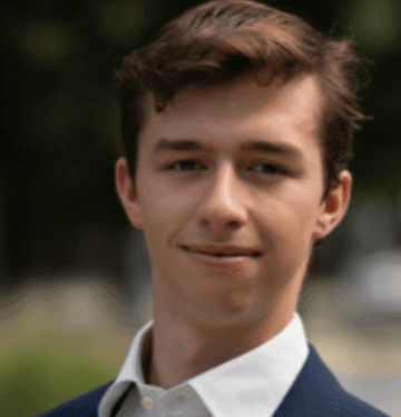 Man wearing a white collared shirt and dark blazer standing outdoors with a blurred natural background.