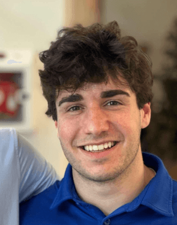 Person with curly hair wearing a blue shirt, standing indoors with a hand around their shoulder.