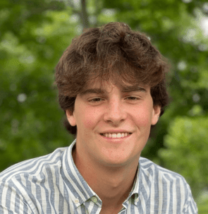 Person with curly brown hair wearing a striped shirt outdoors with green foliage in the background.
